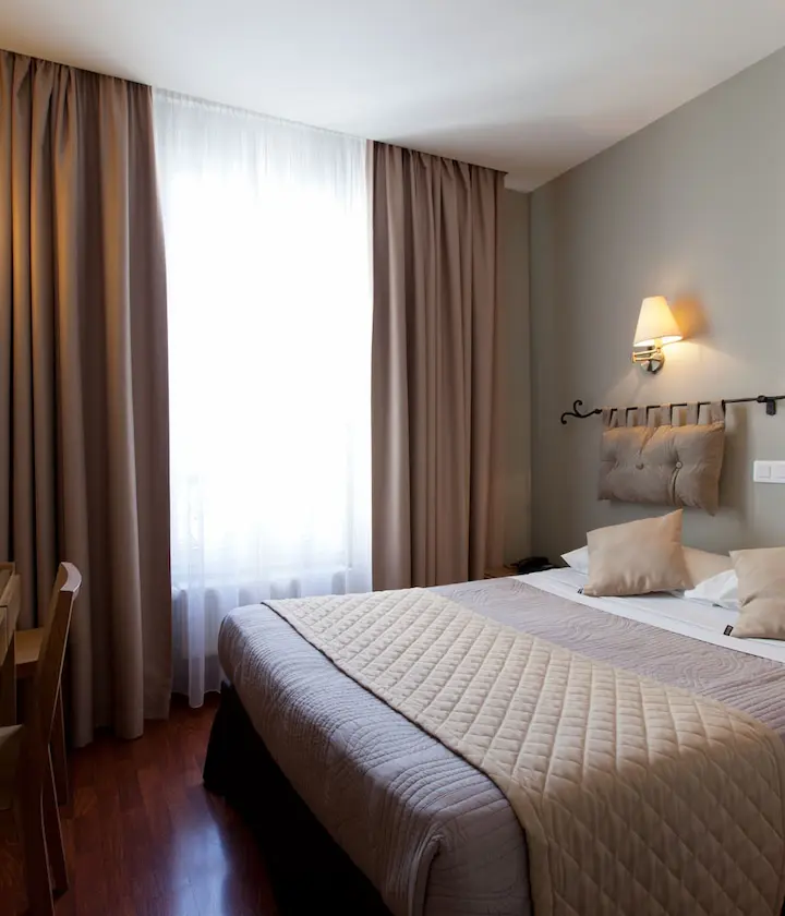 Chambre d'hôtel lumineuse avec lit double, rideaux beiges et applique murale.