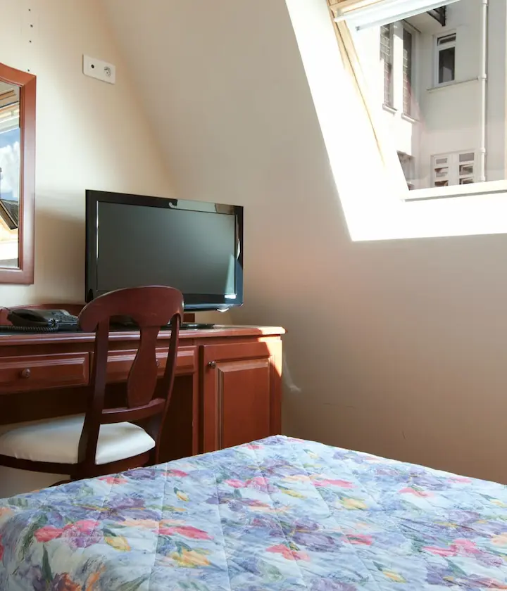 Chambre avec bureau, télé et fenêtre éclairée, lit à la housse colorée.