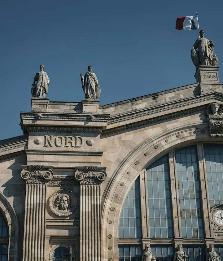 Façade de la Gare du Nord avec statues et drapeau français sous un ciel bleu.