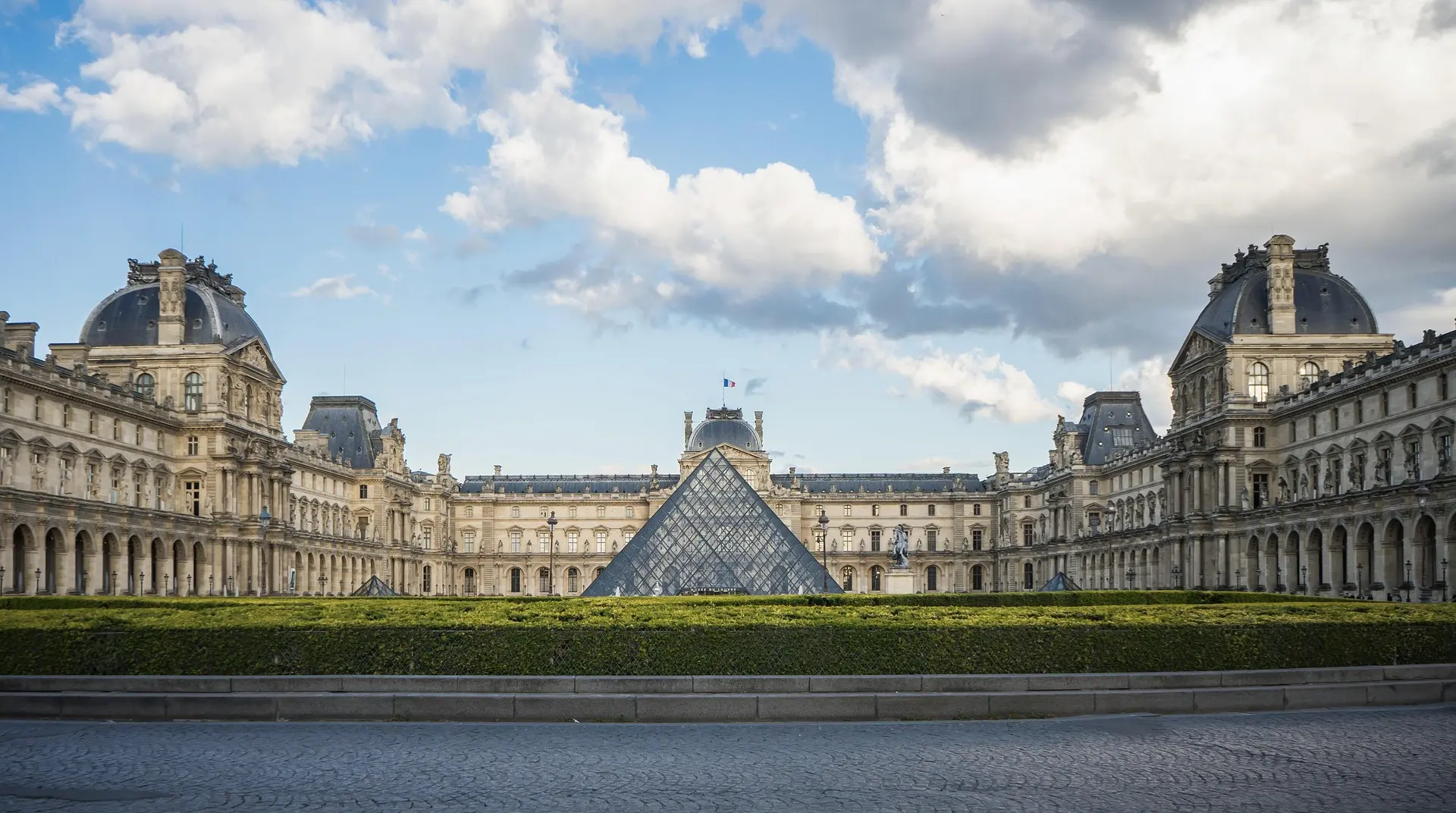Pyramide de verre du Louvre devant le bâtiment historique sous un ciel partiellement nuageux.