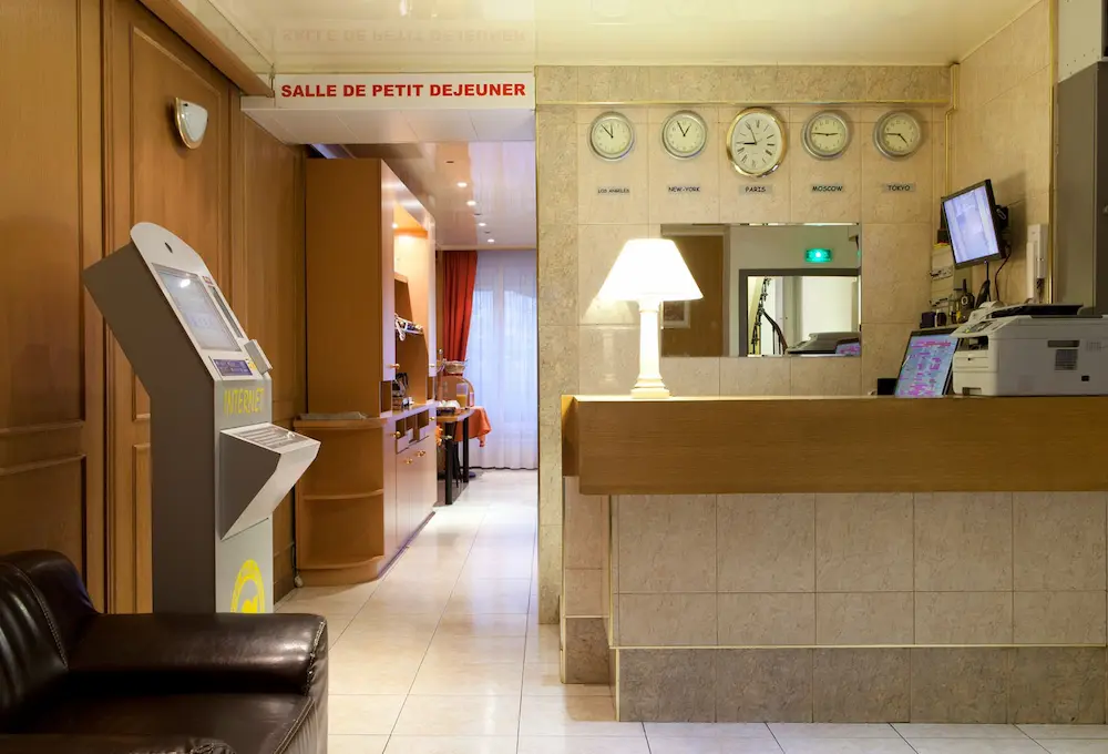 Bureau d'accueil d'hôtel avec lampes, horloges et borne Internet, salle de petit déjeuner en arrière.