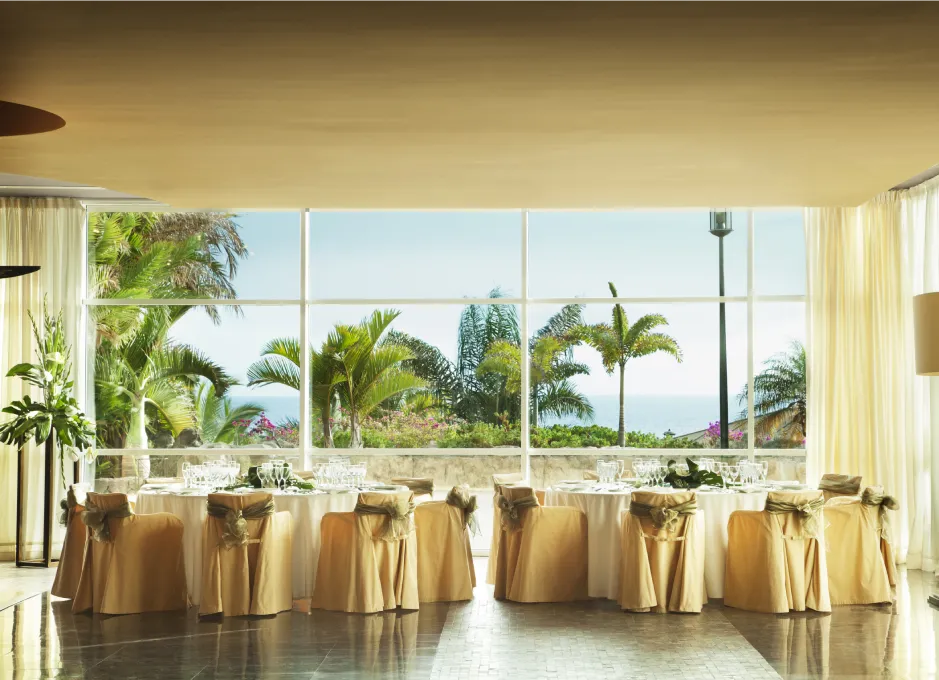 Sala elegante con mesas decoradas y vista a jardín con palmeras.