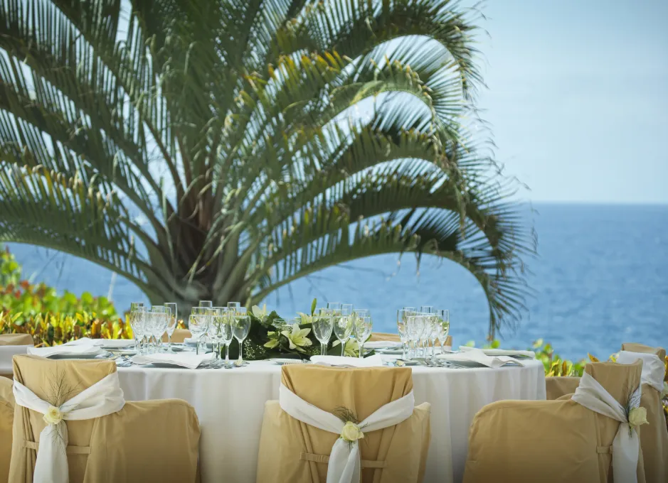 Mesa elegante al aire libre con copas, frente a palmera y mar.