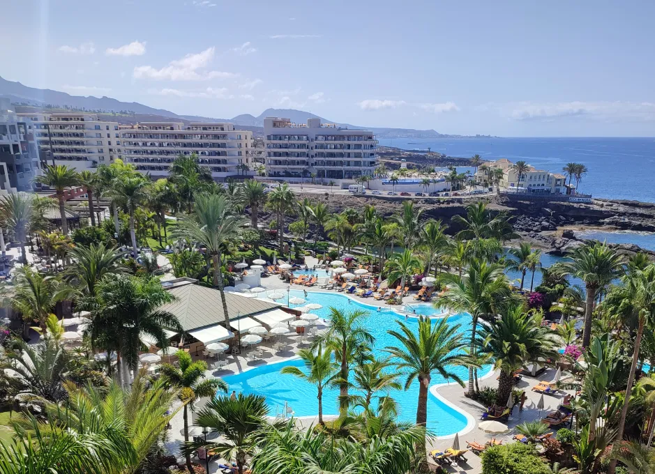 Vista aérea de un complejo turístico con piscina, palmeras y edificios cerca del mar.