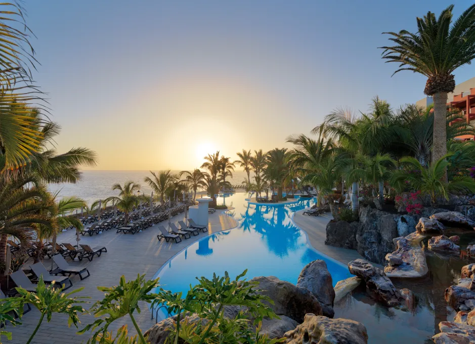 Piscina al amanecer con palmeras y tumbonas, reflejando la luz del sol en un entorno tropical.