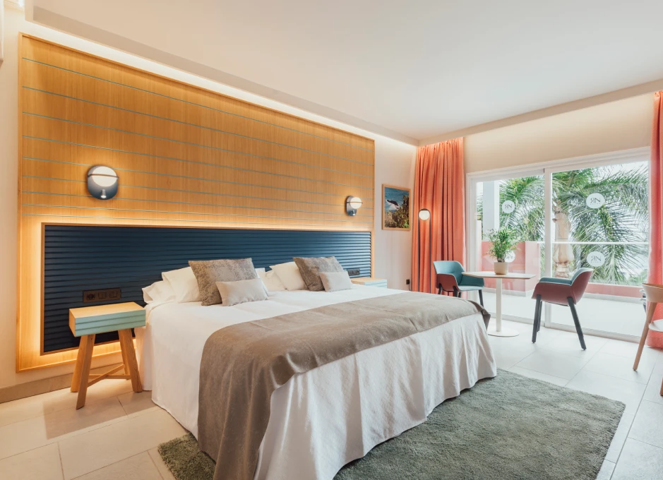 Habitación luminosa con cama grande, mesa y sillas, cortinas naranjas y ventana con vista a palmeras.