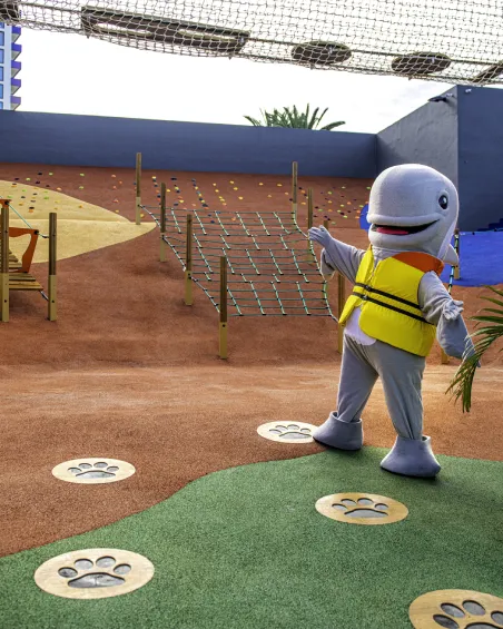 Mascota disfrazada de delfín en un área de juegos con redes y huellas en el suelo.