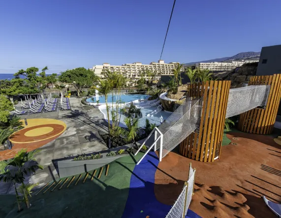 Zona de juegos y piscina al aire libre con edificaciones al fondo bajo un cielo despejado.