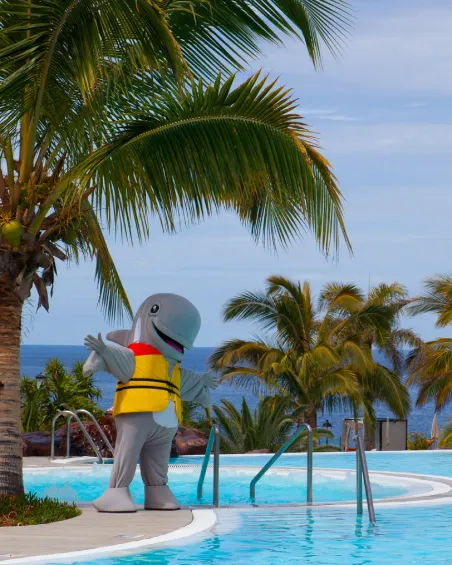 Traje de delfín junto a piscina tropical, rodeado de palmeras y vista al mar.