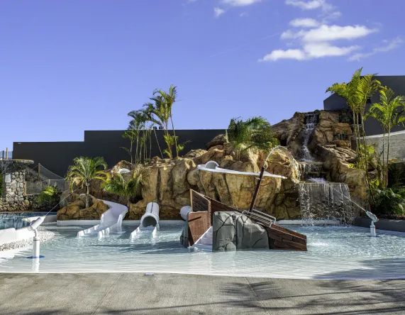 Piscina con toboganes y cascadas rodeada de palmeras y rocas bajo un cielo azul.