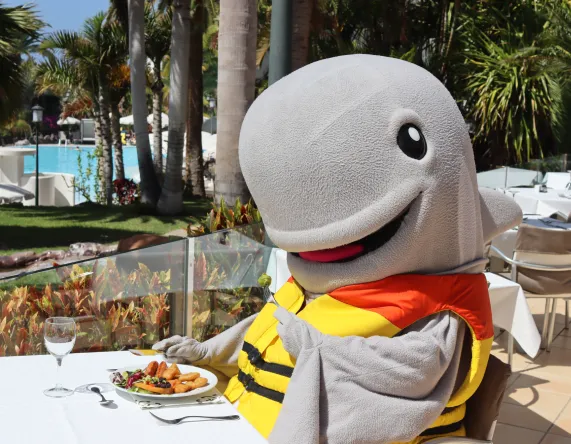 Personaje disfrazado de tiburón comiendo en una mesa al aire libre junto a la piscina.