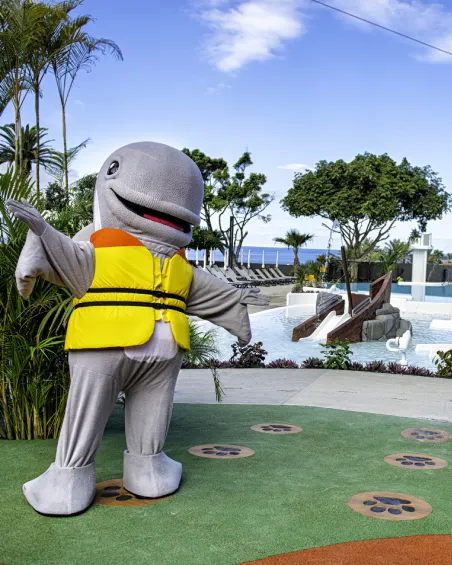 Personaje disfrazado de delfín con chaleco amarillo, frente a una piscina y árboles.