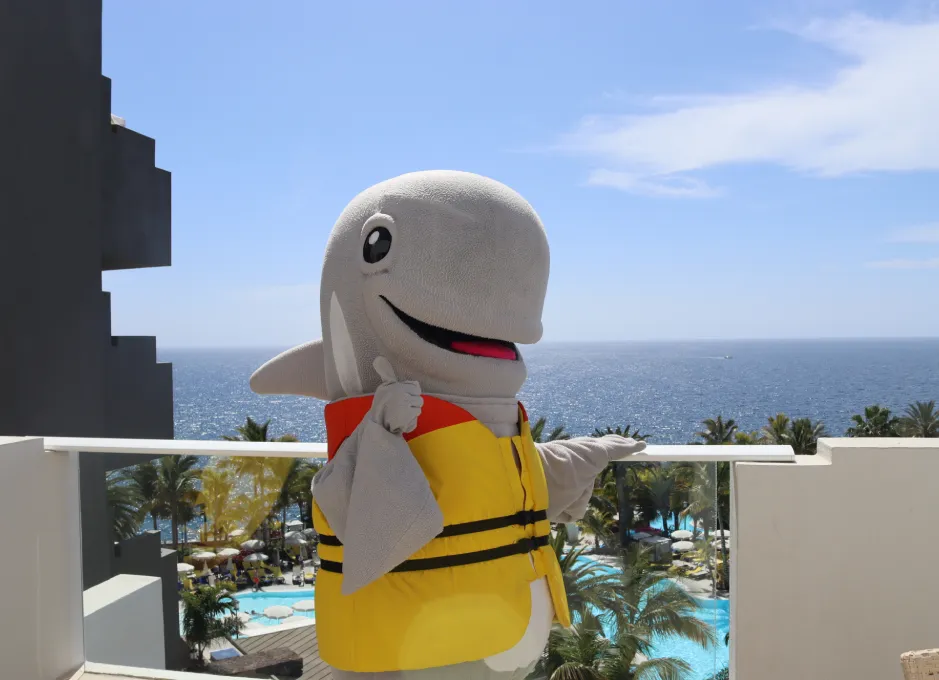 Mascota de delfín con chaleco, sonriendo frente al mar desde un balcón soleado.