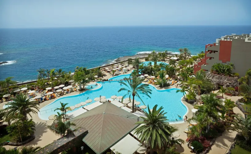 Vista de una piscina con palmeras y el océano de fondo en un complejo turístico.