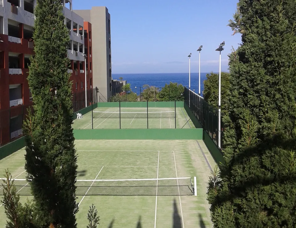 Cancha de tenis vacía entre edificios y árboles, con vista al mar al fondo.