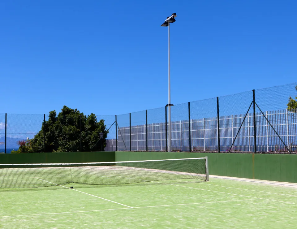 Cancha de tenis al aire libre con cielo despejado y cercada, rodeada de arbustos y luces de poste.