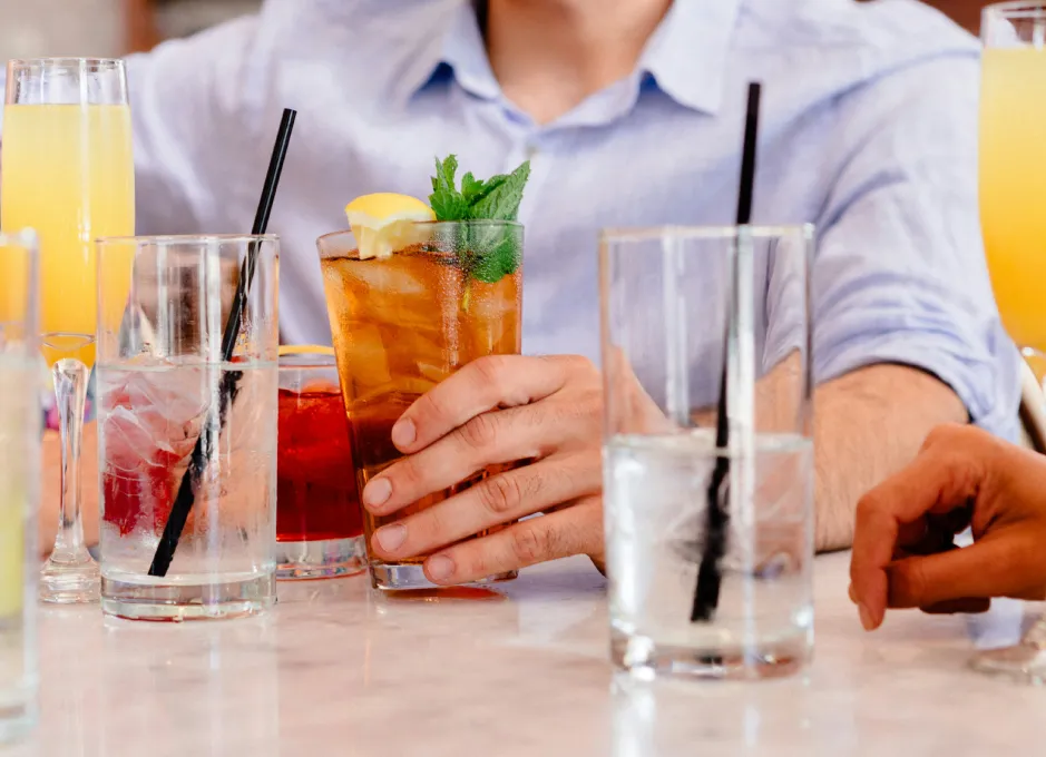 Persona sosteniendo un vaso con bebida de té helado y menta, rodeada de otros vasos en una mesa.