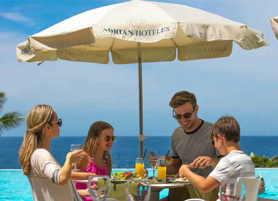 Familia disfrutando de una comida al aire libre bajo una sombrilla junto a una piscina y el mar.