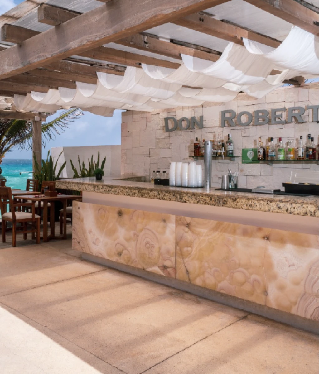 Bar al aire libre con decoración elegante y vista al mar, con mesas y sillas de madera, cubierto por un techo de vigas y tela.