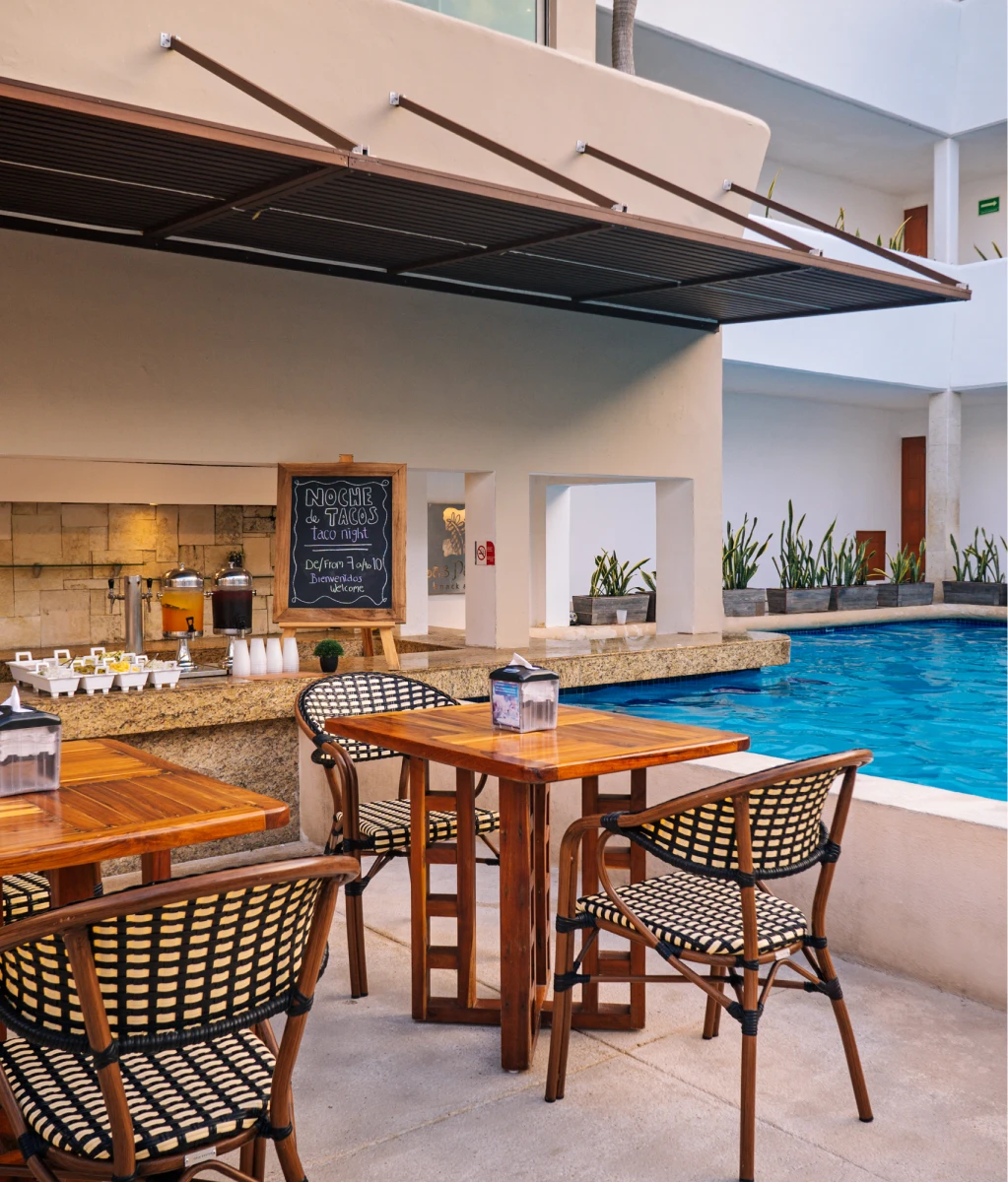 Área de comedor al aire libre junto a una piscina, con mesas de madera, sillas tejidas y un cartel de "Noche de Tacos".