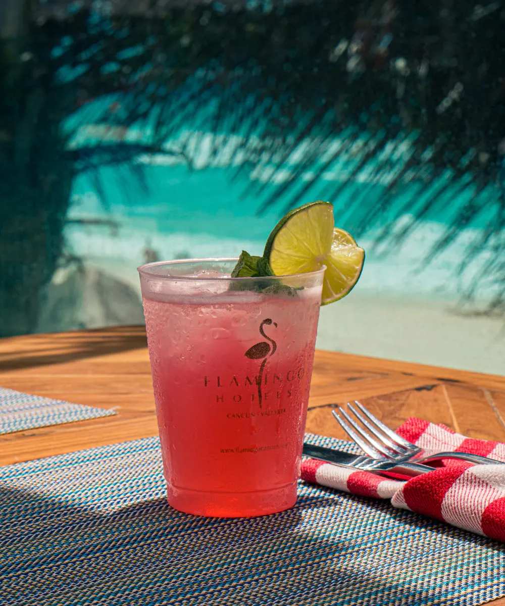 Cóctel rosa con limón en vaso de plástico sobre una mesa junto a cubiertos, fondo de playa y palmeras.