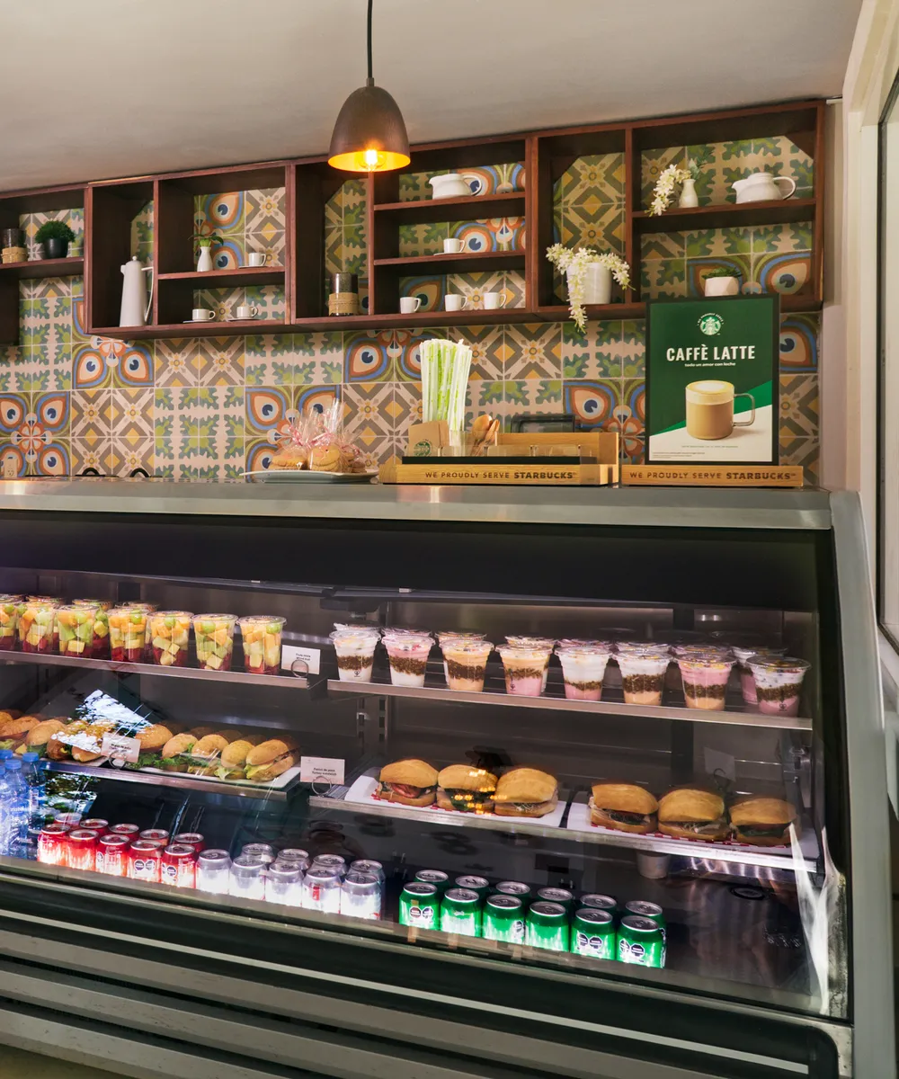 Vitrina de cafetería con frutas, postres, sándwiches y bebidas, frente a estantes decorativos y una pared colorida.