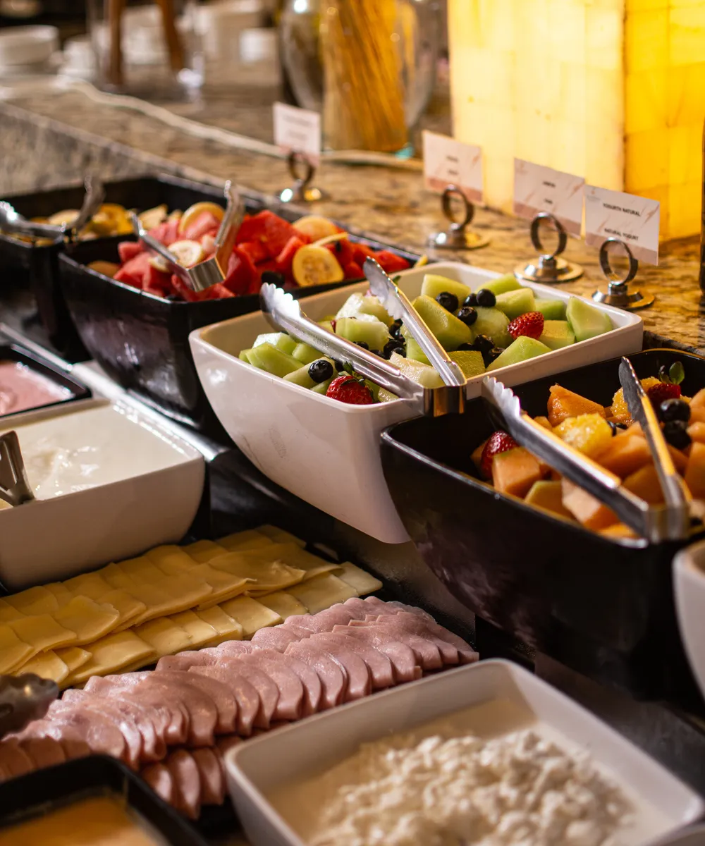 Bandejas de buffet con frutas variadas, quesos, carnes frías y salsas en una mesa de mármol.