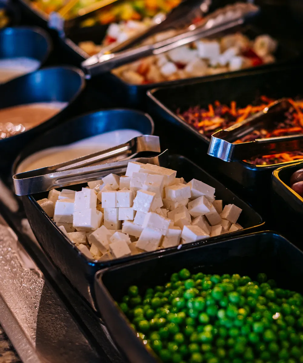 Bandejas de buffet con tofu en cubos, guisantes y ensaladas variadas con pinzas metálicas.