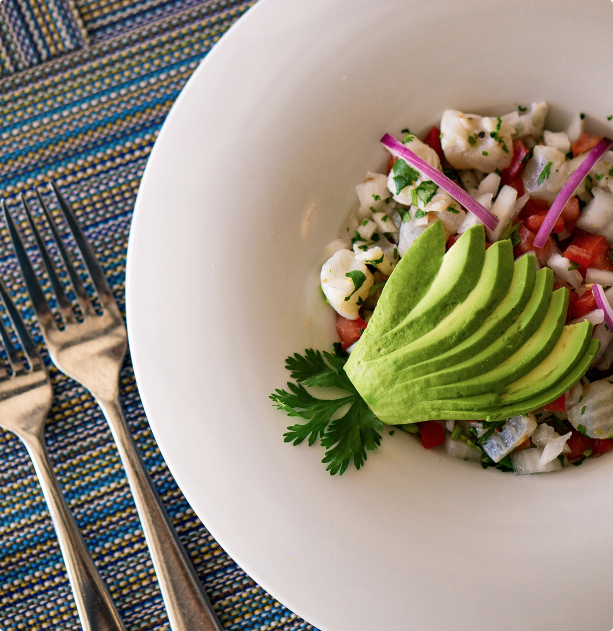 Plato de ceviche con rodajas de aguacate y perejil decorativo, sobre un mantel azul, junto a un par de tenedores.