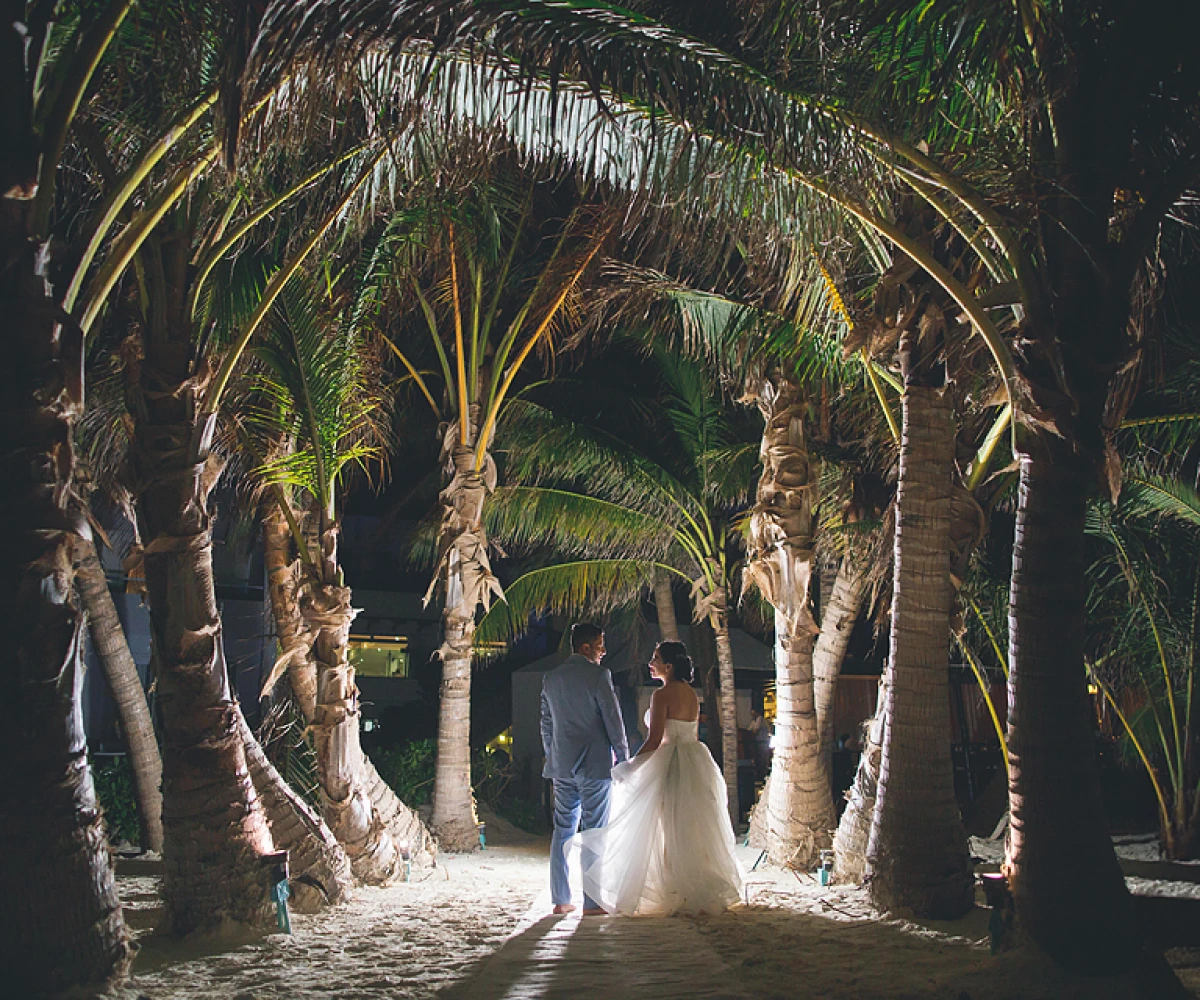 Pareja en su boda, caminando descalzos entre palmeras iluminadas.