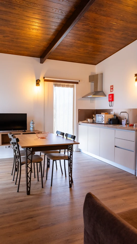 Cozinha integrada com mesa, cadeiras, TV e bancada sob teto de madeira.