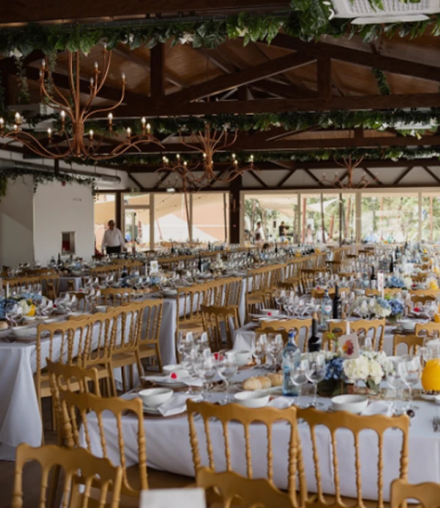 Salão de festas decorado com mesas arrumadas, cadeiras douradas e lustres elegantes.