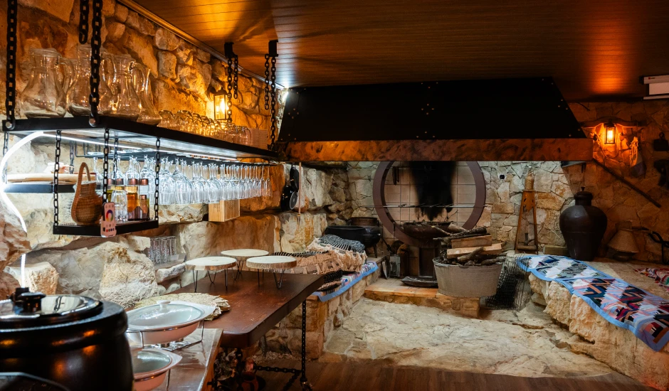 Adega rústica com paredes de pedra, copos pendurados, forno e decoração tradicional.