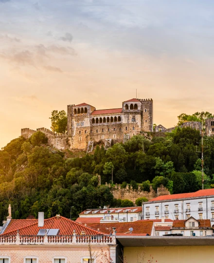 Castelo em uma colina coberta de árvores com casas ao redor, ao entardecer.