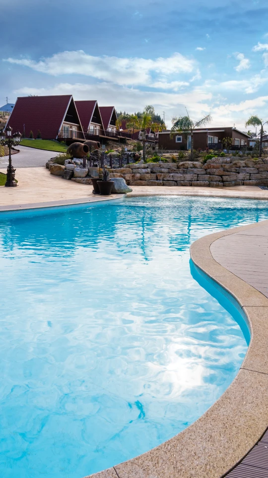 Piscina em forma orgânica com chalés triangulares ao fundo, céu azul e nuvens.