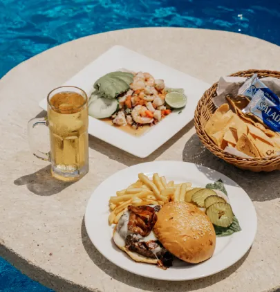 Platos de comida y cerveza junto a una piscina: hamburguesa, camarones, papas y nachos.
