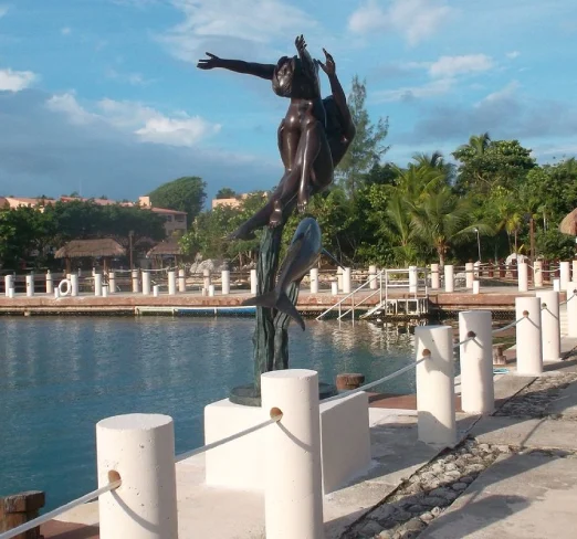 Estatua de bronce de un hombre con un delfín en un muelle rodeado de agua y vegetación.