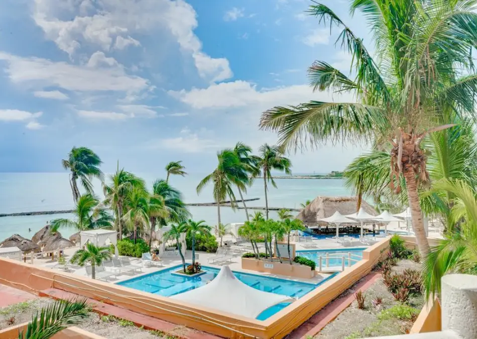 Vista de una piscina y palmeras junto a la playa, bajo un cielo parcialmente nublado.