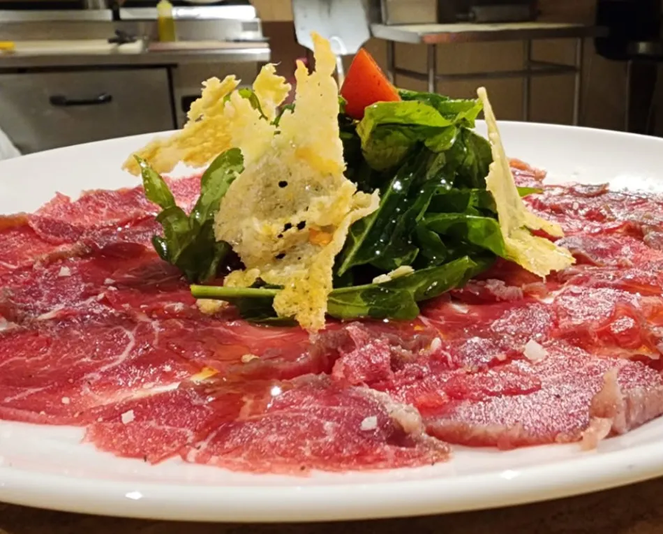 Carpaccio de carne con rúcula y trozos de queso crujiente sobre un plato blanco.