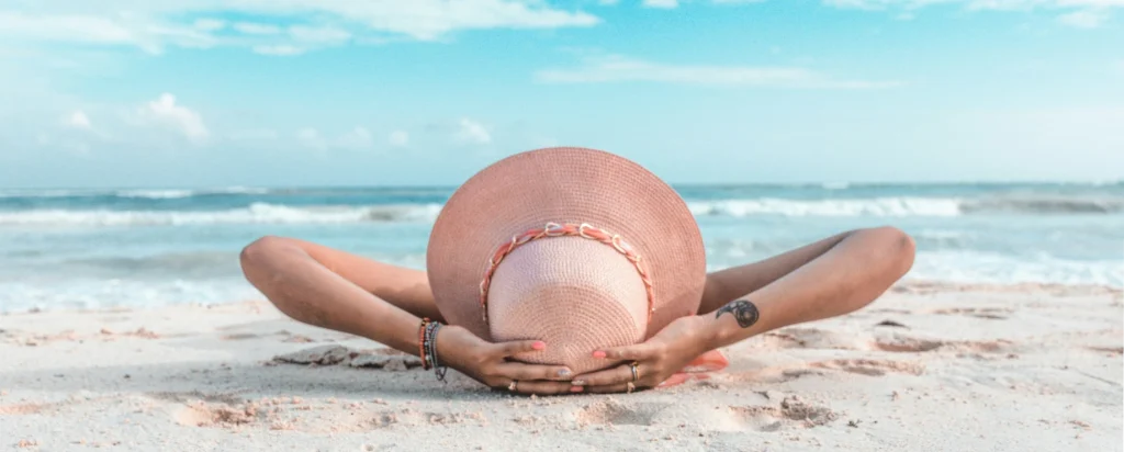 Mujer tumbada en la playa boca abajo con sombrero rosa, mirando al mar y cielo azul claro.