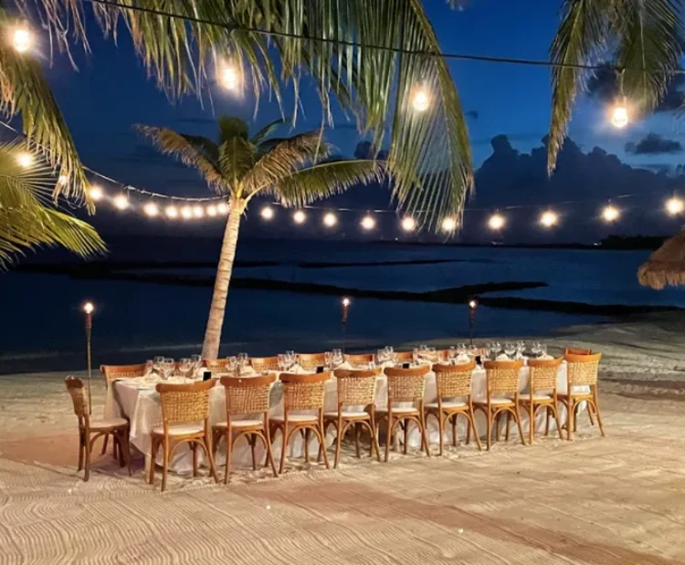Mesa al aire libre con luces colgantes en una playa, rodeada de palmeras al atardecer.