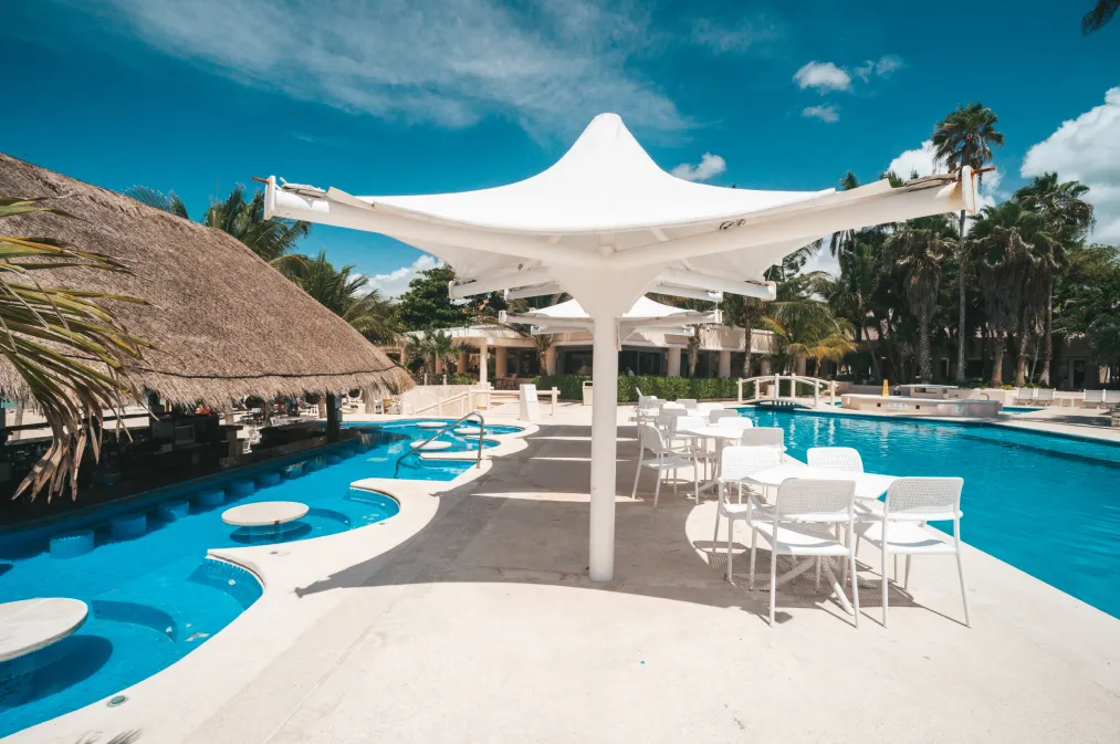 Piscina con mesas y sillas blancas al aire libre, rodeada de palmeras y cielo azul despejado.