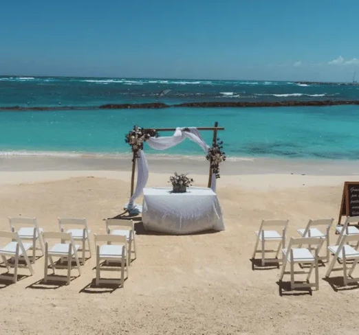 Ceremonia de boda en la playa con sillas blancas y altar frente al mar azul.