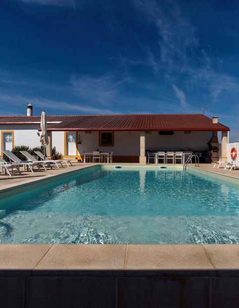 Piscina ao ar livre com espreguiçadeiras e casa pintada de branco ao fundo. Céu azul claro.