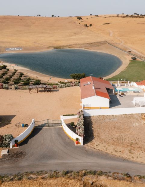Casa com telhado vermelho ao lado de lagoa azul, cercada de campos dourados e flores.