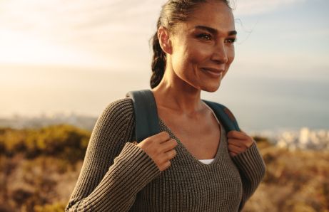 Mulher sorrindo com mochila nas costas, ao ar livre, com paisagem desfocada ao fundo.