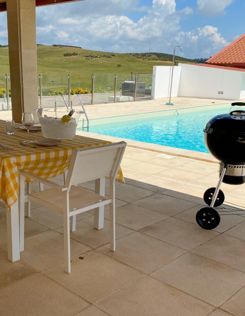 Área externa com mesa posta, churrasqueira e piscina ao fundo em dia ensolarado.
