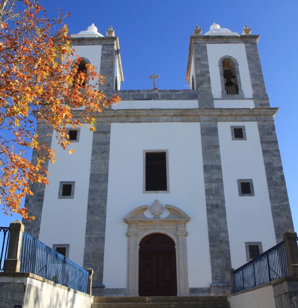 Fachada de uma igreja branca com duas torres, céu azul e árvore com folhas outonais ao lado.