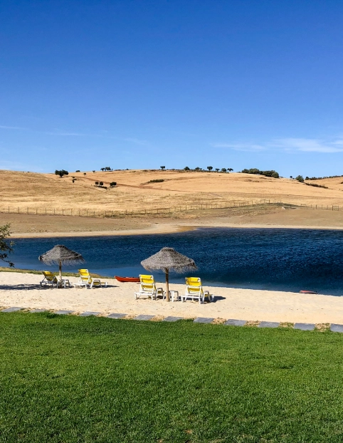 Praia com cadeiras e guarda-sóis de palha, lago e colinas ao fundo.