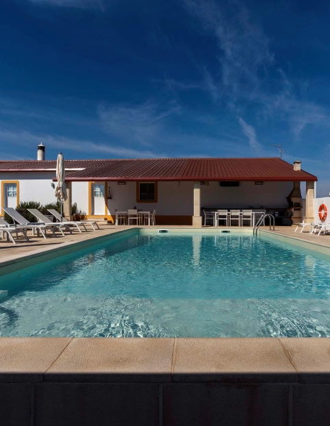 Piscina ao ar livre em frente a uma casa com espreguiçadeiras e céu azul.